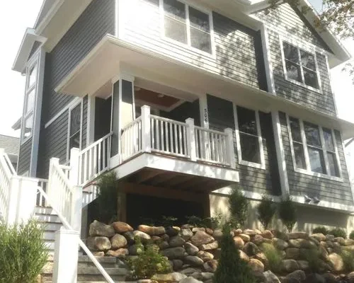 A similar two-story home with gray siding, white trim, and a covered porch. The home is positioned on a hillside with a retaining wall made of large rocks.