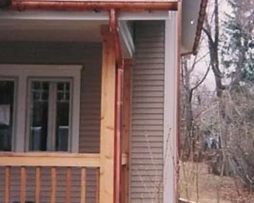 An image of the exterior of a wooden house with a covered porch, a gabled roof, and various architectural details.